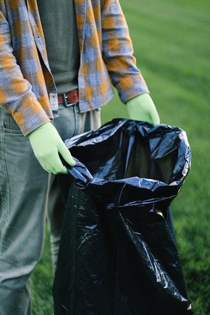 A person standing outdoors on a grassy area is holding open a large black rubbish bag lined with a silver inner layer. The individual is wearing a checked shirt with shades of blue, gray, and mustard 